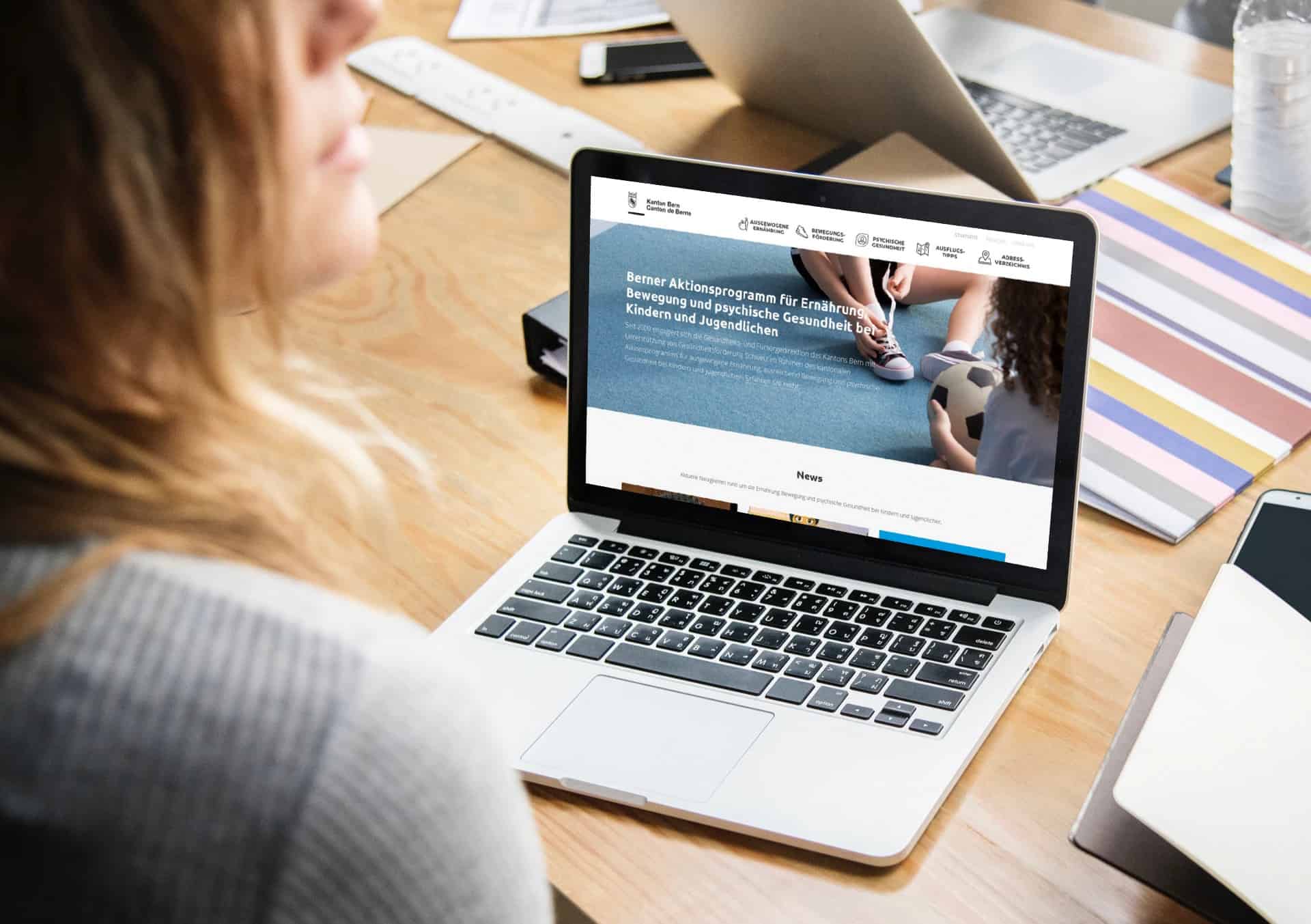 A person sits at a desk in Basel with a laptop on which a German-language WordPress website is displayed, surrounded by documents, a telephone and a cup of water. The site presents a health or sports program for children and young people.