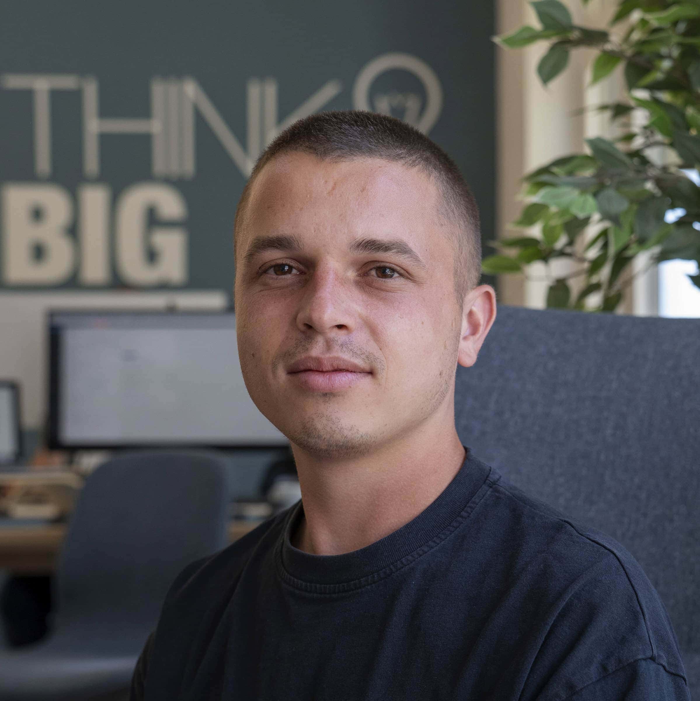 Ein Mann mit kurzen Haaren und gestutztem Bart, der ein dunkles T-Shirt trägt, sitzt in einem Büro mit Computern, einer Pflanze und dem Schriftzug "THINK BIG" an der Wand, der auf einen kreativen Arbeitsbereich der WordPress Agentur hinweist.